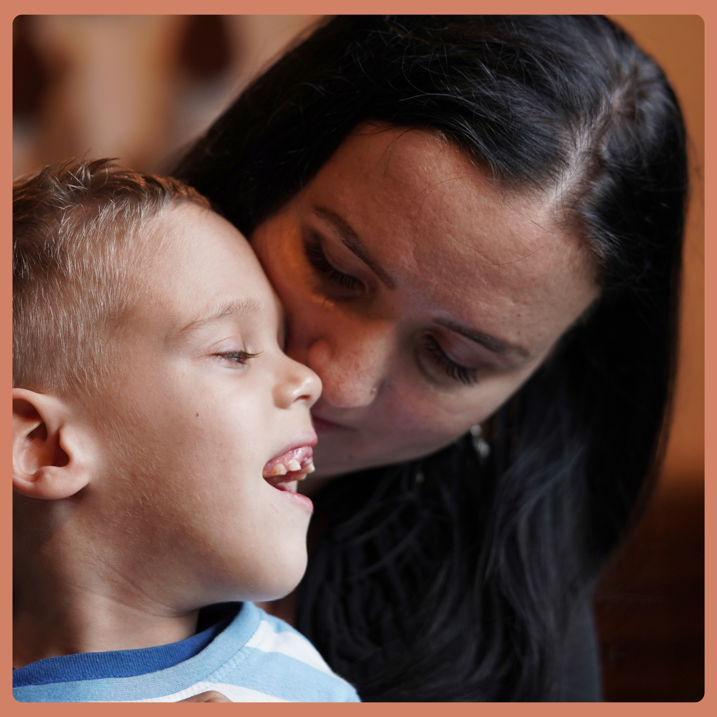 A mom is snuggled into the neck of her neurodivergent son, who is smiling - his missing two front teeth are prominent with his big smile.