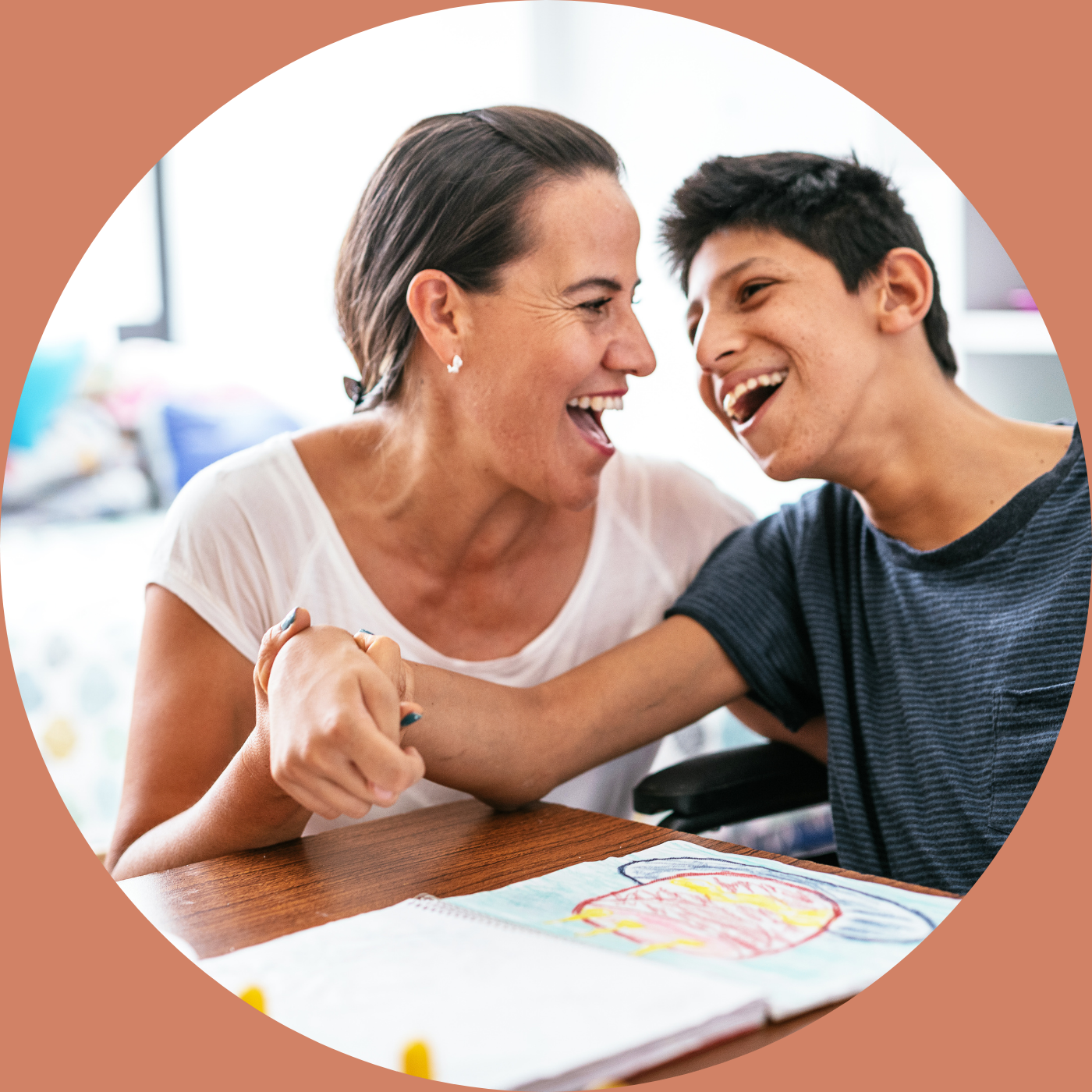 A woman holds her neurodivergent son's hand. They are sitting at the table with a drawing in front of them. Both are laughing.